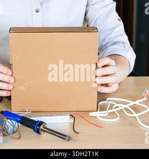 Close-up of person's hands lifting a plain cardboard box above a table scattered with wires, soldering iron, and power supplies in a DIY setup. Stock Photo