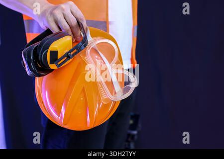Worker gripping bright orange hard hat featuring black earmuffs and transparent goggles on dark background for safety concept. Stock Photo