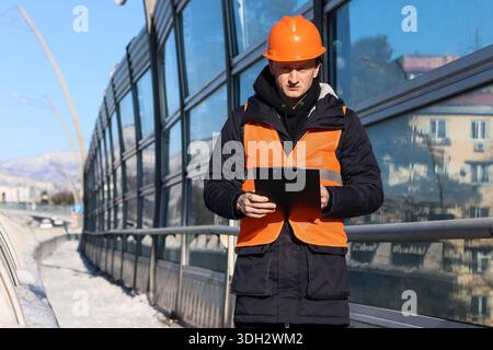 A man in orange safety attire reviews paperwork outdoors on a snowy elevated structure. Stock Photo