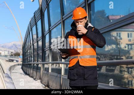 Male engineer in safety gear drinks hot beverage and checks documents outdoors in snowy urban setting Stock Photo