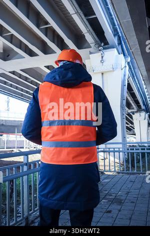 Close-up back shot of safety worker in reflective gear inspecting bridge underside. Stock Photo