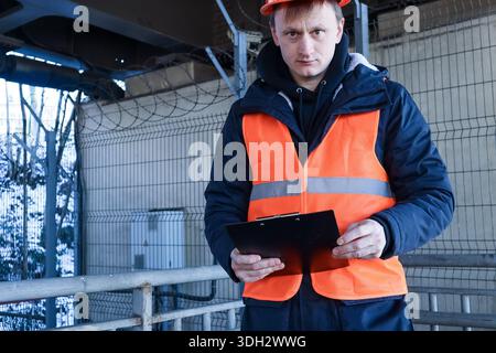 Front view of focused male worker in orange reflective vest and helmet standing with clipboard at secure fenced site Stock Photo
