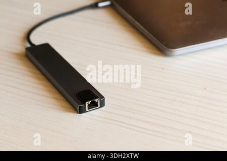 Sleek black USB-C to Ethernet adapter plugged into a laptop, enabling wired network connectivity on a light wooden desk Stock Photo