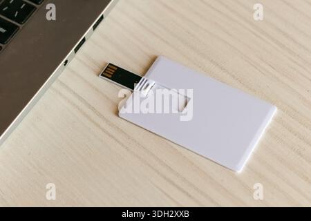 Slim credit card-shaped USB drive with extended flash stick placed adjacent to a silver laptop on a wooden desk, offering slimline data storage soluti Stock Photo