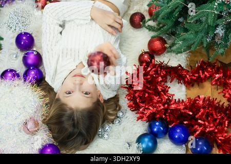 Kid in light clothing extends hand to glossy ball on white plush area amid colorful baubles and garlands Stock Photo