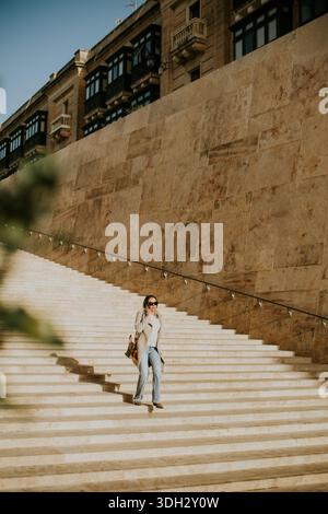 A woman walks down a set of stone steps in Malta. The setting is bright with clear skies. Buildings can be seen in the background. The woman carries a Stock Photo