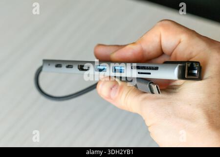Detailed shot of a hand holding a gray multi-function USB-C hub with Ethernet, USB, and HDMI ports connected by cable Stock Photo