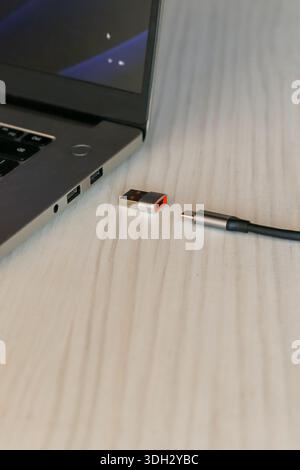 Laptop with glowing blue screen, a USB dongle adapter with orange trim, and USB-C cable arranged on a light wooden surface for device charging. Stock Photo