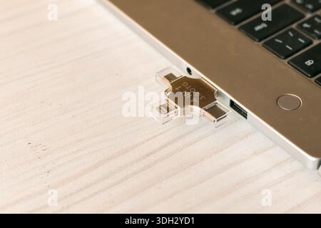 Side angle of a slim silver laptop featuring a plugged-in multi-connector USB flash drive on a wooden table, ideal for cross-device data synchronizati Stock Photo