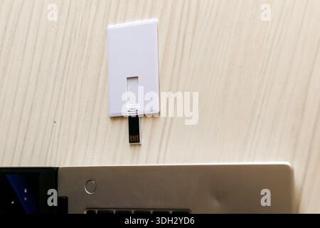 Flat white USB card drive featuring a protruding black connector, positioned next to a silver laptop's keyboard on a light wooden table for quick use. Stock Photo