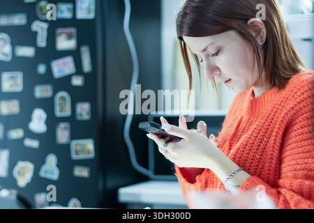 Close-up of a focused young woman holding and scrolling on her smartphone near a wall decorated with various photos and stickers in a modern interior. Stock Photo