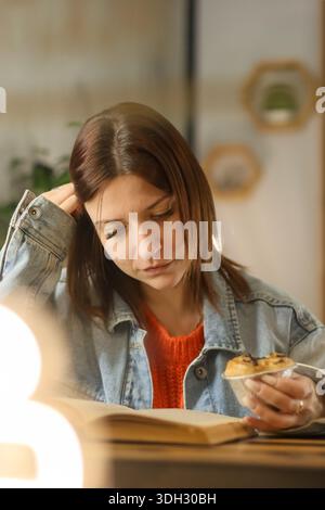 Young woman in denim jacket reading a book at cafe table, holding muffin, thoughtful expression, cozy atmosphere Stock Photo
