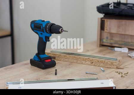 A blue battery-powered drill placed on a table with wooden boards, metal rails, screws, and assembly items nearby. Stock Photo