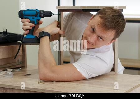 Concentrated young man in white shirt leans under light wood frame, using blue cordless drill to tighten screw from below on table. Stock Photo