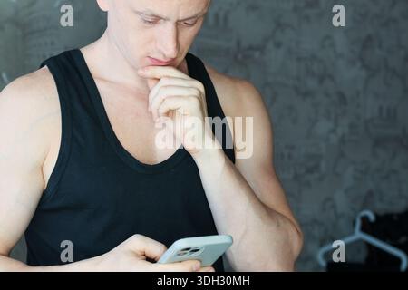 Fit male in sleeveless shirt touching chin contemplatively, holding mobile device indoors Stock Photo