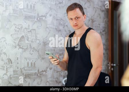 Casual young man in tank top standing indoors, holding smartphone in hand with decorative wallpaper behind. Stock Photo