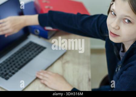 Close-up of preteen girl in blue top extending arm to point at laptop display, with hand on keyboard and colorful folders stacked beside. Stock Photo