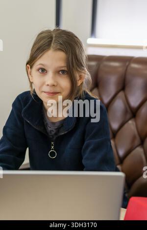 Close-up of a preteen girl in navy jacket smiling softly at the viewer, seated at a desk with a laptop and red notebook visible. Stock Photo