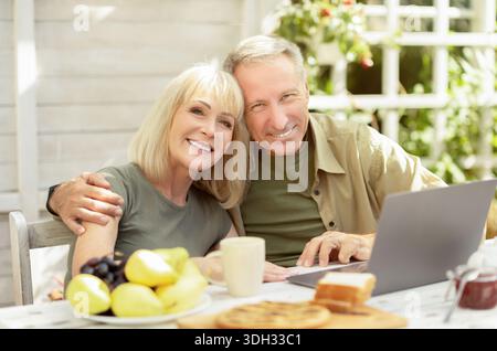 Happy senior couple using laptop computer outdoors, sitting at home terrace and smiling to camera Stock Photo