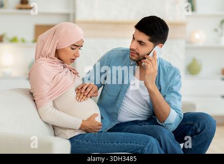 Pregnant arab woman having prenatal contractions, worried husband calling doctor, using mobile phone, home interior Stock Photo