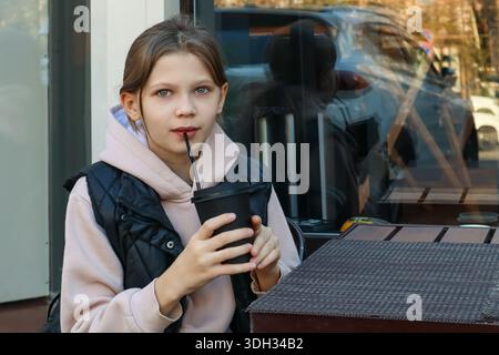 Close-up of a girl with blue eyes pursing lips around a straw from a disposable cup at an al fresco dining spot in city environment. Stock Photo