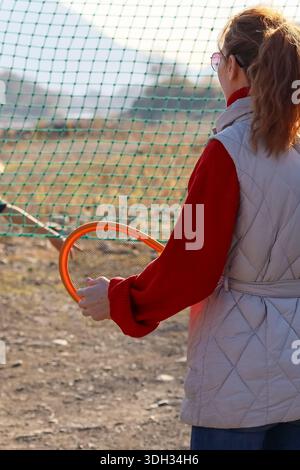 Rear view of female player gripping colorful racket by green net against mountainous backdrop in autumn. Stock Photo