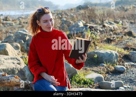 Casual young female sitting on log by river, enjoying outdoor reading in sunny weather Stock Photo