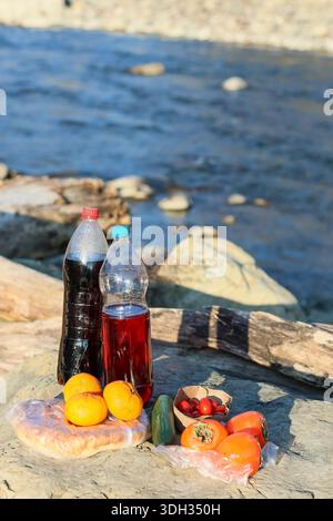 Assortment of picnic items like soft drink, cordial, ripe persimmons, cucumber, cherry tomatoes, bread on driftwood and rocks beside serene river flow Stock Photo