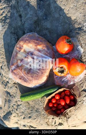 Close-up of wrapped flatbread, persimmons, cucumber, and cherry tomatoes in paper bowl on textured rock surface, highlighting natural textures. Stock Photo
