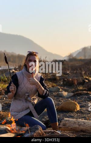 Cozy autumn evening by the river: woman in puffer vest roasts marshmallows over campfire amid scenic mountains and golden light. Stock Photo
