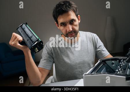 Young man holding empty toner cartridge from laser printer, looking directly at camera with frustrated expression while attempting to fix office Stock Photo