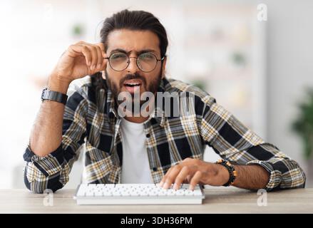 Confused middle-eastern man programmer sitting at worktable, touching glasses Stock Photo