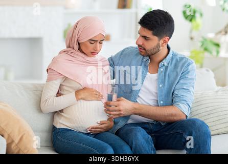 Care and support during pregnancy. Muslim expecting woman feeling sick, man giving her glass of water, siting on sofa Stock Photo