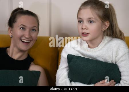 Happy mother and daughter relaxing on a mustard yellow sofa at home, sharing a joyful moment and engaging in conversation while spending quality time Stock Photo