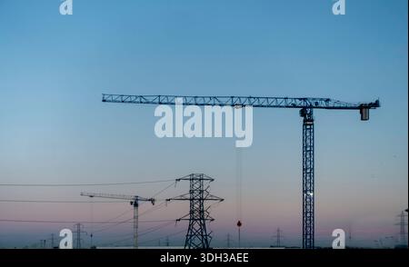 Industrial cranes and electricity pylons silhouetted against a dusk sky at sunset. Infrastructure and construction equipment in a twilight landscape. Stock Photo