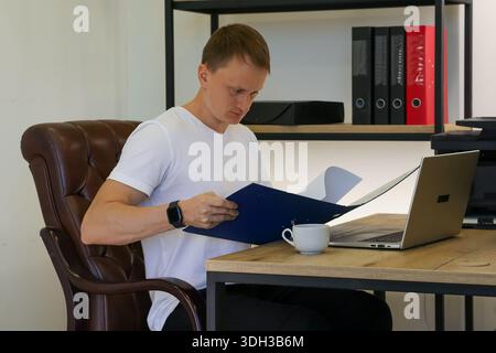 Attentive young man scrutinizes open blue folder with papers in quiet office, laptop on desk accompanied by simple coffee mug. Stock Photo