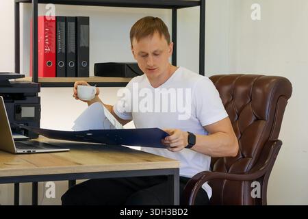 Seated man holds white cup, gazing puzzled at open blue folder with documents on desk, surrounded by office equipment. Stock Photo
