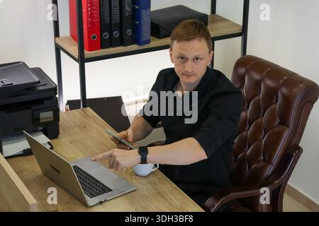 Focused male in black shirt extends arm pointing ahead, holding phone, with laptop and cup on wooden desk. Stock Photo