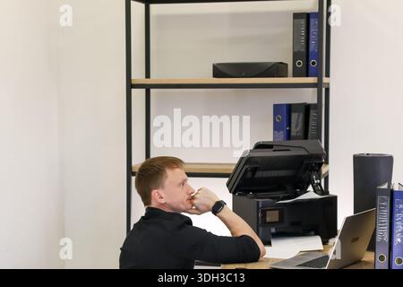 Thoughtful worker in black clothing contemplates printer issue while seated at desk Stock Photo