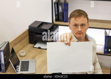 Worker in shirt offering empty white document towards viewer amid office furniture and devices Stock Photo