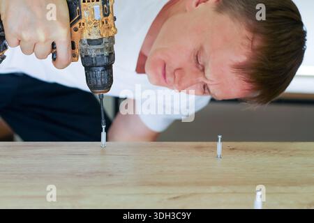 Profile view of male with serious expression using yellow power drill to insert dowel into wood plank in bright indoor workspace. Stock Photo