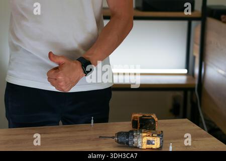 Satisfied craftsman showing thumbs up gesture next to power drills and fasteners on wooden surface Stock Photo