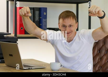 Joyful young man in white shirt pumps both fists in victory while smiling at open laptop on wooden desk with cup nearby. Stock Photo