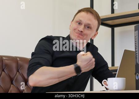 Delighted businessman grins ear-to-ear with fist pumped in achievement, wristwatch visible, sips from white coffee cup, laptop on desk, amid stylish o Stock Photo