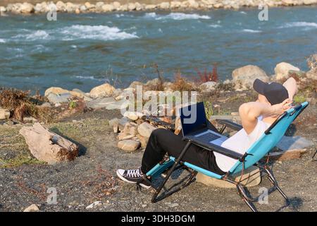 Lounging comfortably in a blue recliner chair near the riverbank rocks, a young man stretches with laptop on his lap in a serene natural setting. Stock Photo
