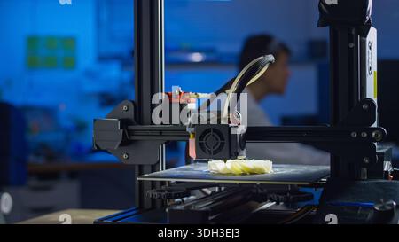 A professional engineer adjusts a 3D printer in a lab while monitoring the printing process for precision. Stock Photo