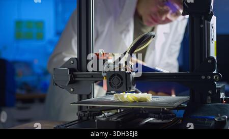 Engineer in lab coat and safety glasses adjusts 3D printer while creating a prototype in a modern lab focused on advanced technology. Stock Photo