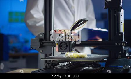 Engineer in lab coat and safety glasses oversees 3D printing process in modern workspace, focusing on precision and technology. Stock Photo