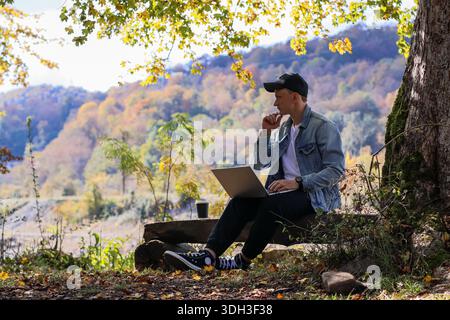 Relaxed man holds cigarette to lips while typing on open laptop on rustic bench in sunny fall woods, coffee steam rising, background of orange foliage Stock Photo