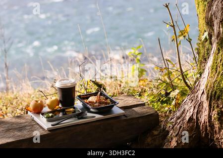 Dark takeaway coffee cup with white lid, black container of grain dish with fork, two apples, small snacks in trays, utensils on silver laptop on wood Stock Photo